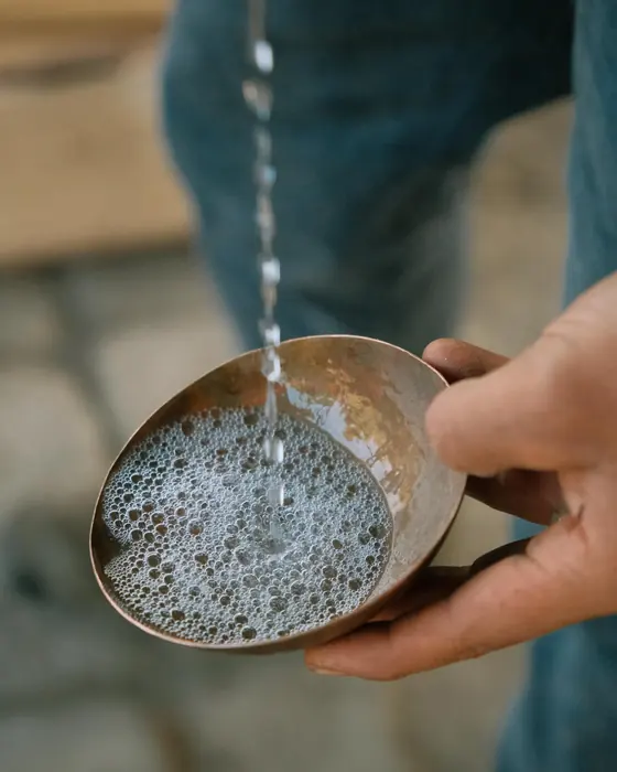 Unas manos sostienen una jícara de metal bajo el hilo de mezcal que cae, con las perlas formándose en la superficie.